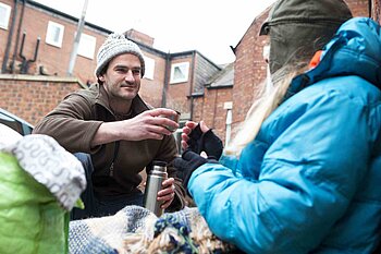 Eine Person reicht einer anderen im Freien einen Becher; beide tragen Winterkleidung