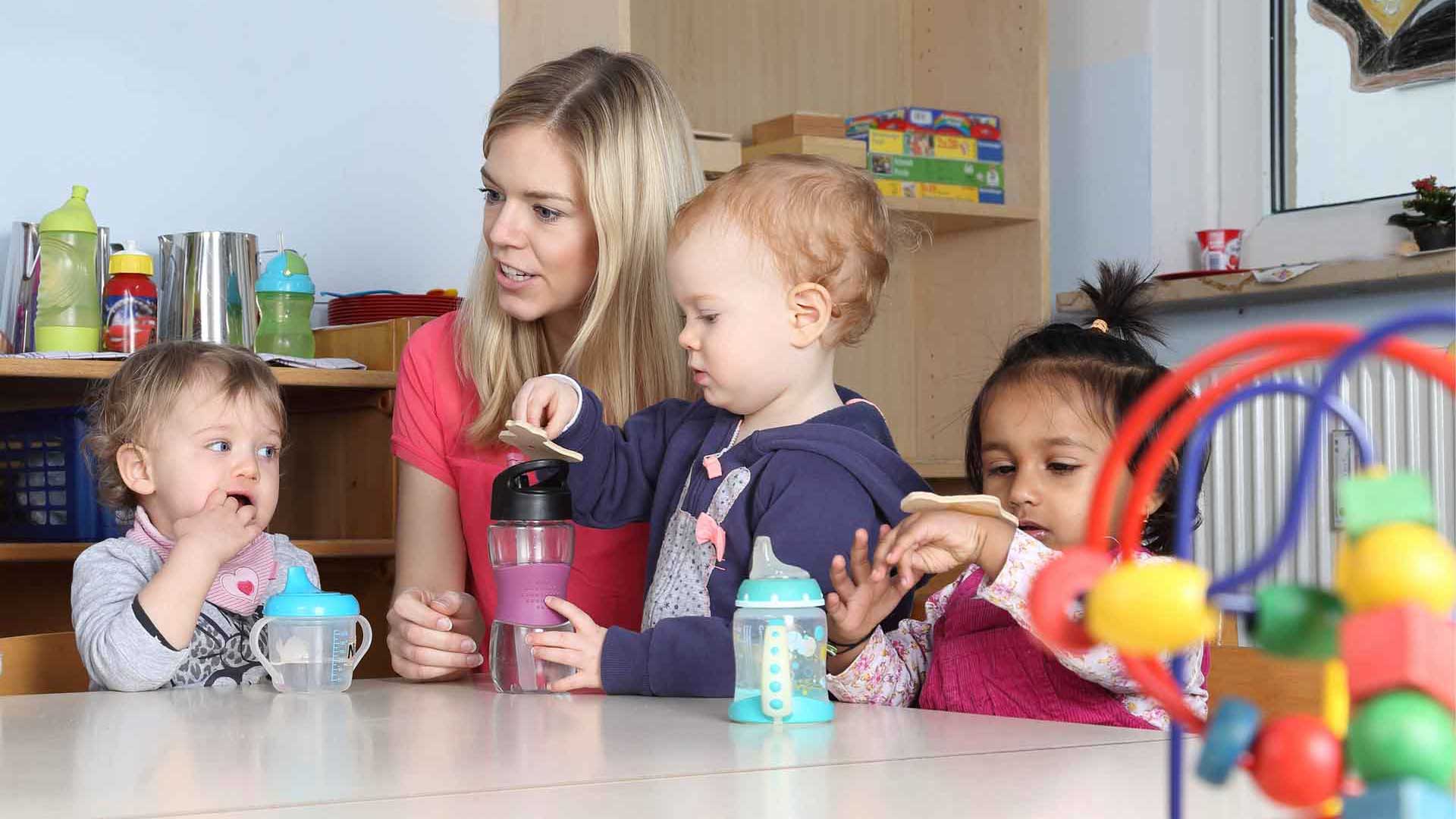 Eine erwachsene Person unterstützt Kleinkinder bei einer gemeinsamen Alltagssituation im Innenraum.