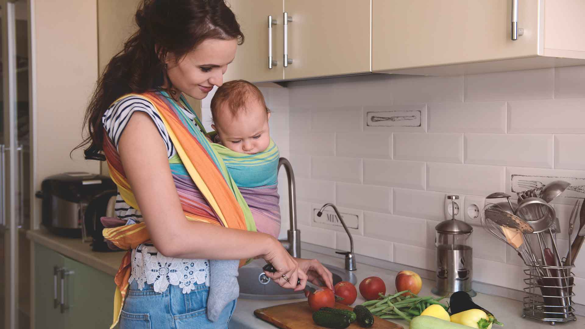 Eine Frau mit Baby im Tragetuch bereitet Essen in der Küche zu.