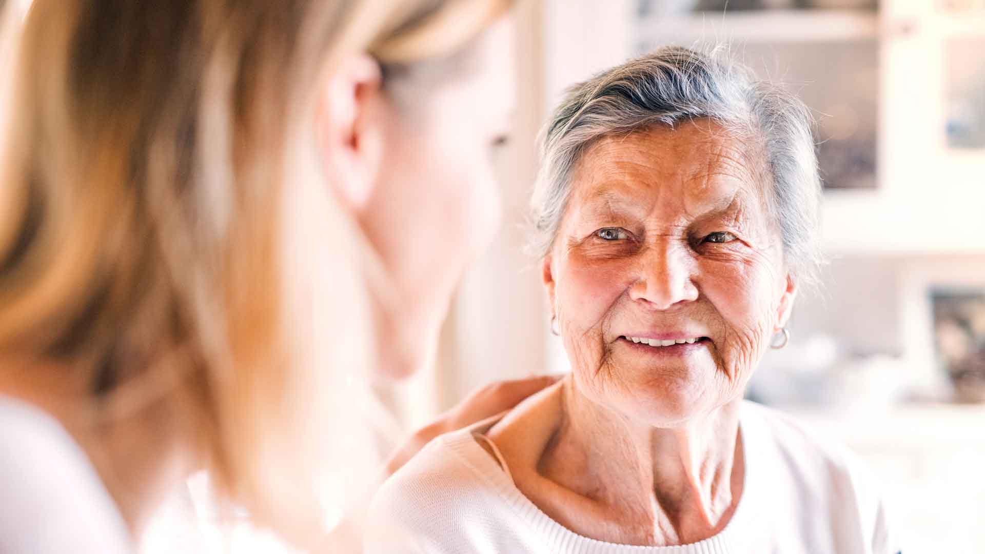 Eine ältere Frau mit grauen Haaren blickt lächelnd eine jüngere Frau an, die unscharf im Vordergrund zu sehen ist.
