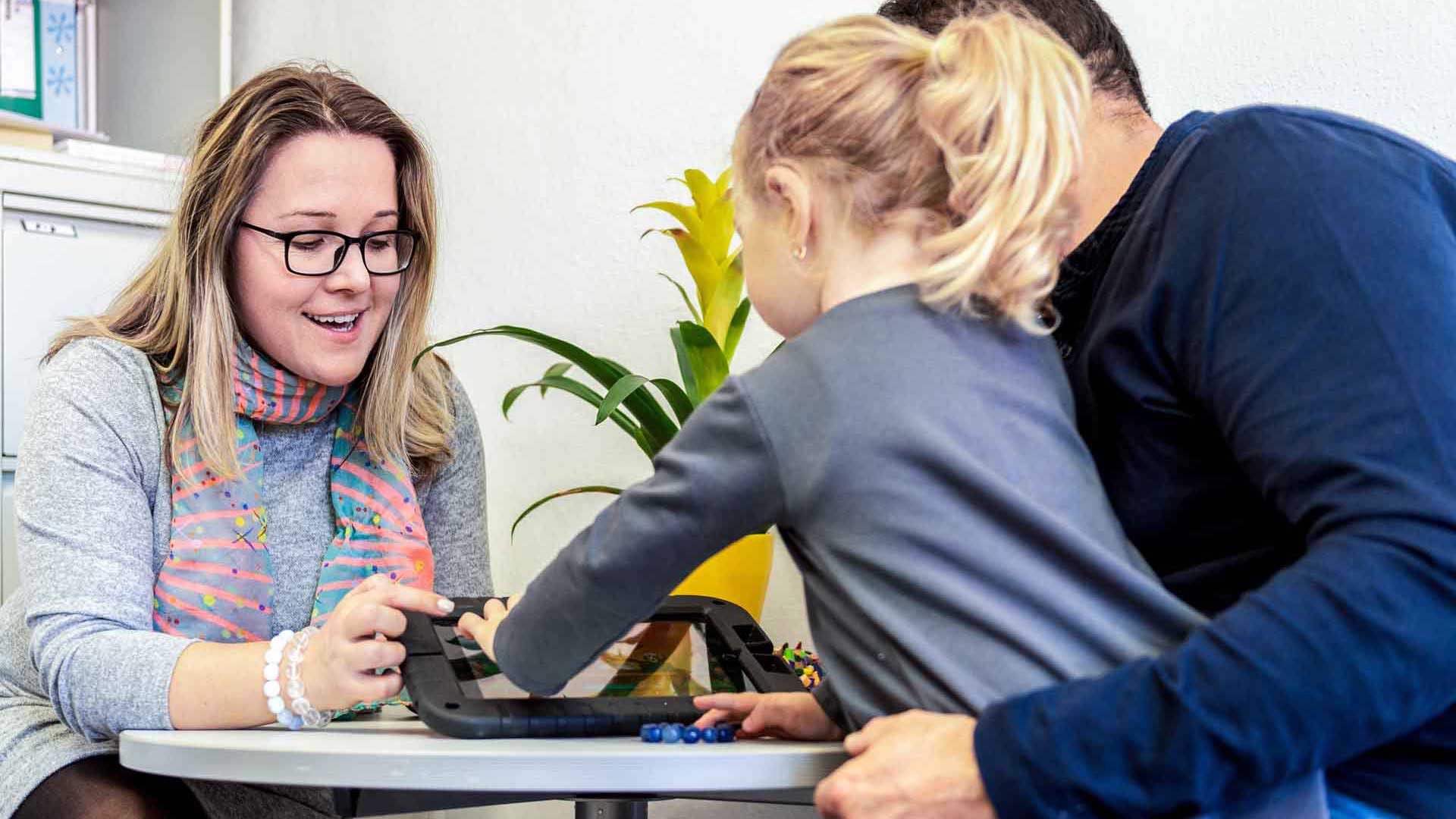 Zwei Erwachsene sitzen mit einem Kind an einem Tisch und zeigen etwas auf einem Tablet.