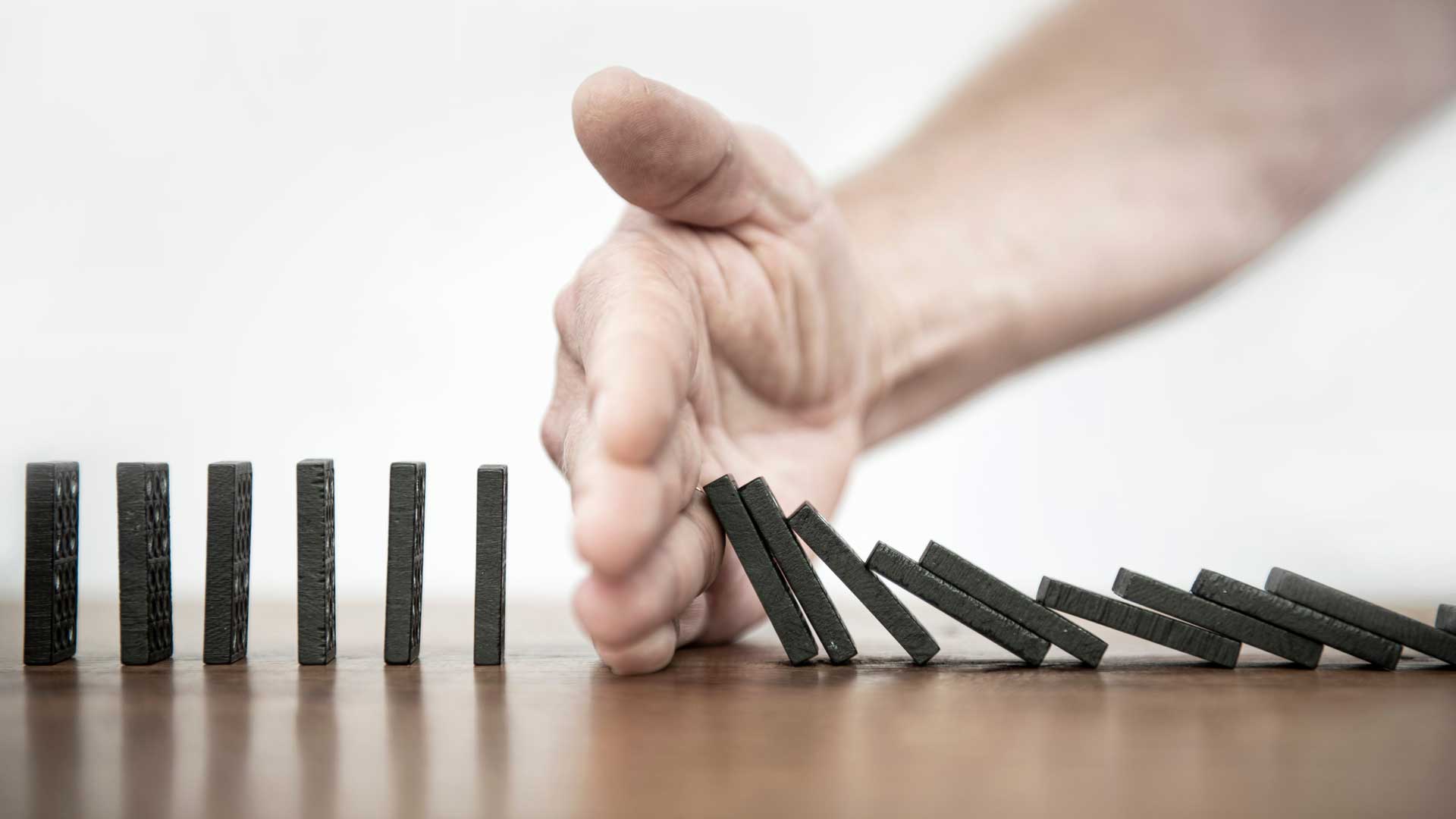 Auf einem Tisch stehen schwarze Dominosteine, die von einer Hand am Weiterfallen gehindert werden.