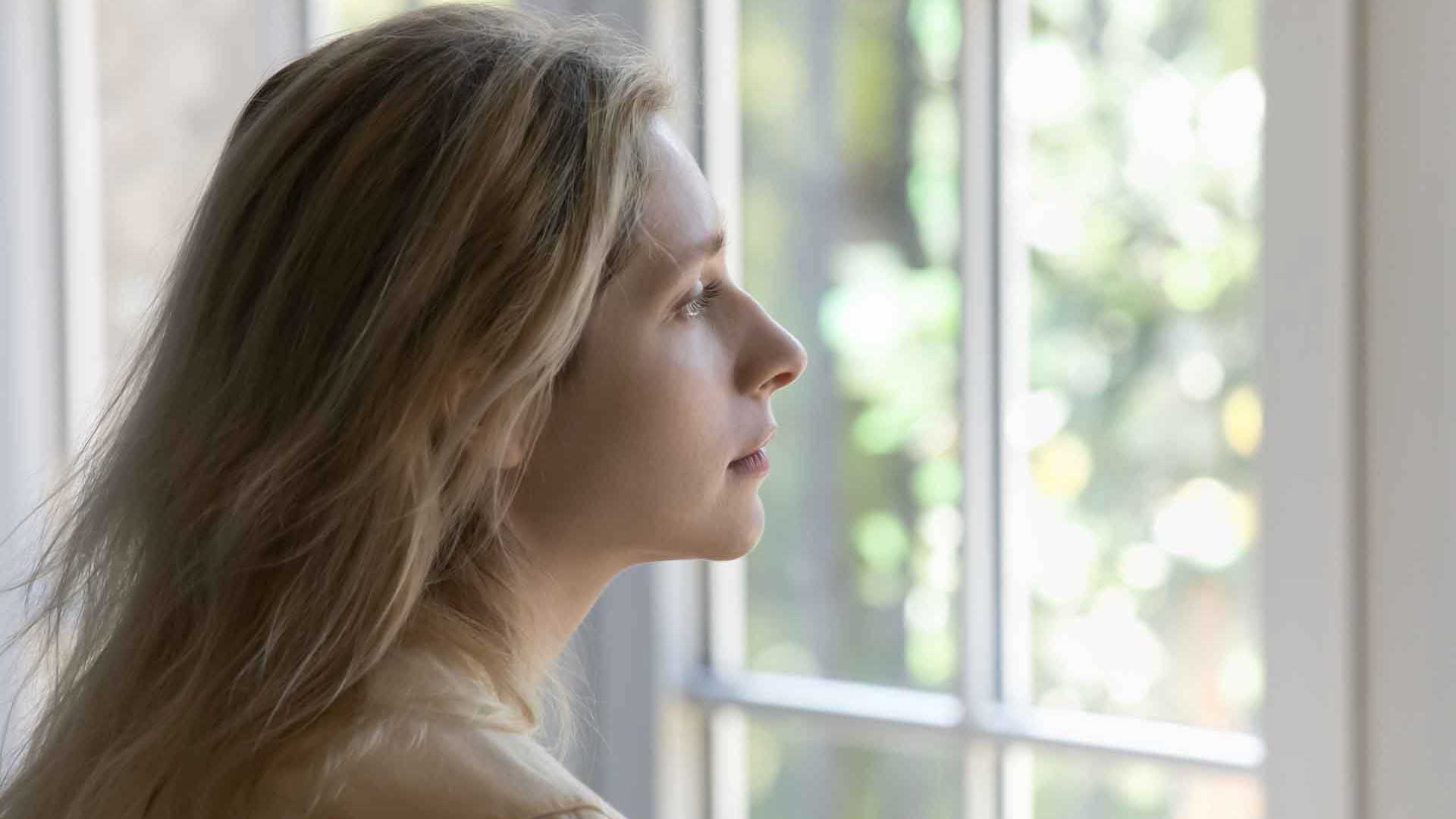 Eine junge Frau im Profil steht an einem Fenster und blickt nach draußen.