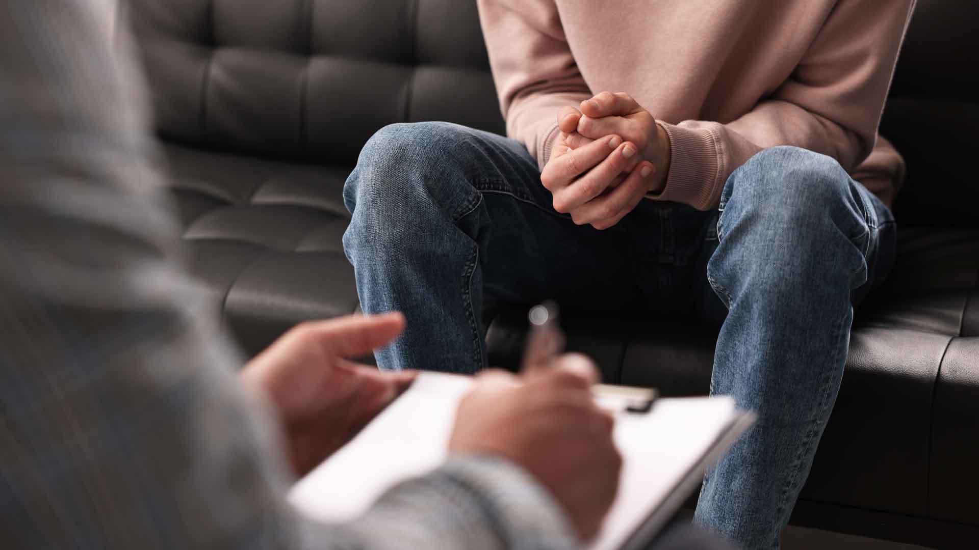 Eine sitzende Person mit verschränkten Händen auf den Knien; im Vordergrund hält eine andere Person ein Klemmbrett.